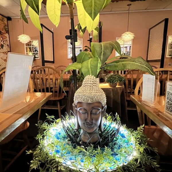 A Buddha statue centerpiece with glowing blue stones and greenery, surrounded by restaurant tables and chairs.